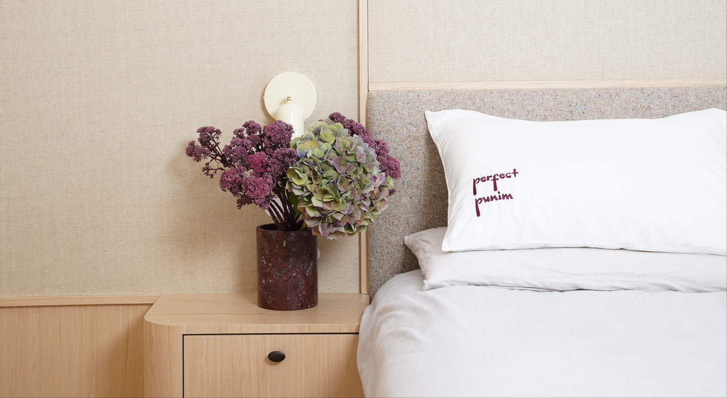 Bedroom with a vase of flowers on a nightstand next to a bed with a branded pillow.