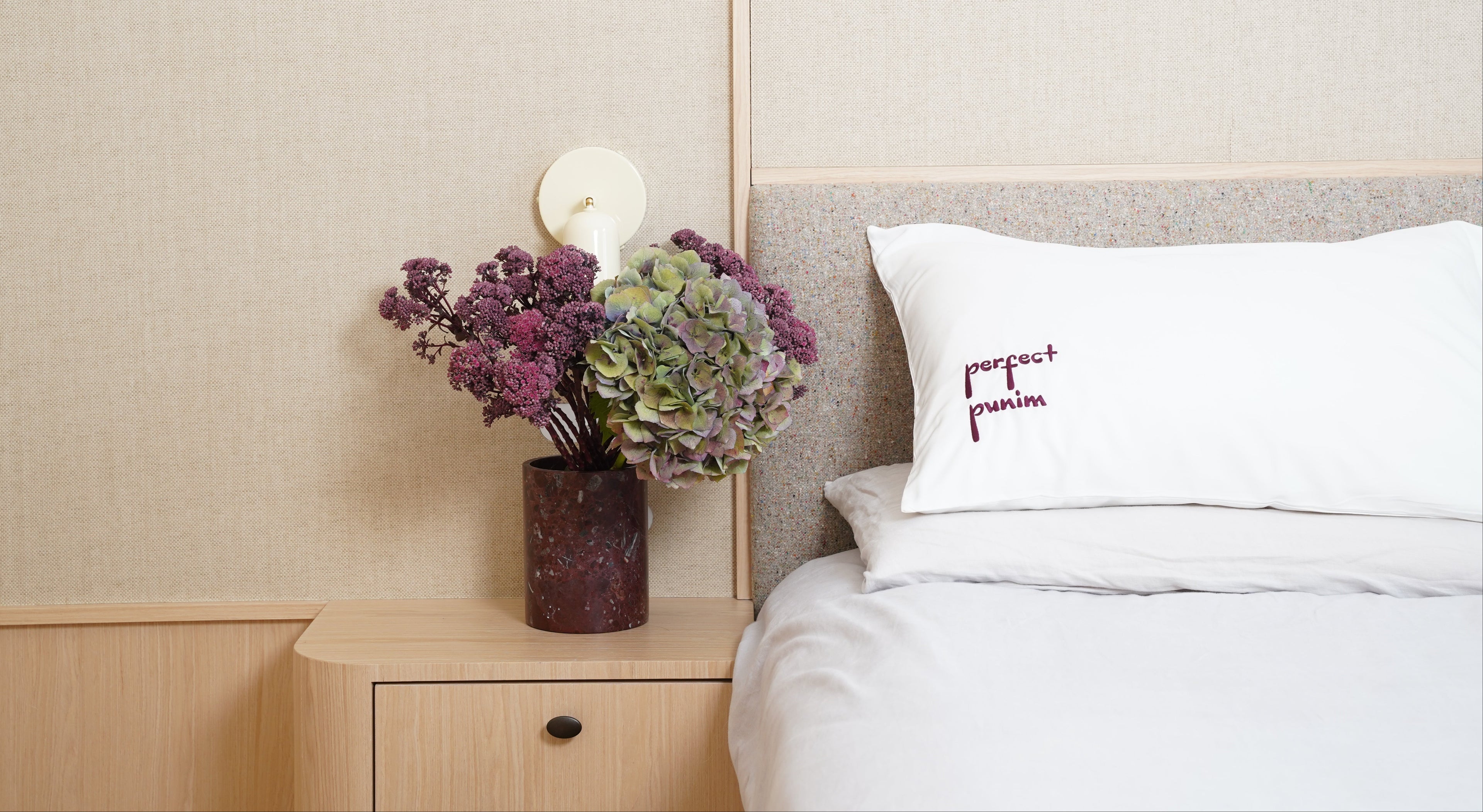 Bedroom with a vase of flowers on a nightstand next to a bed with a branded pillow.